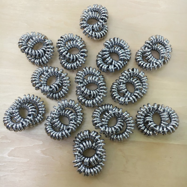 Puede incluir: Un conjunto de coleteros plateados en espiral dispuestos sobre una superficie de madera clara. Los coleteros est&aacute;n hechos de un material met&aacute;lico brillante y enrollados en forma circular. La imagen muestra varios coleteros, lo que sugiere un conjunto o compra al por mayor.