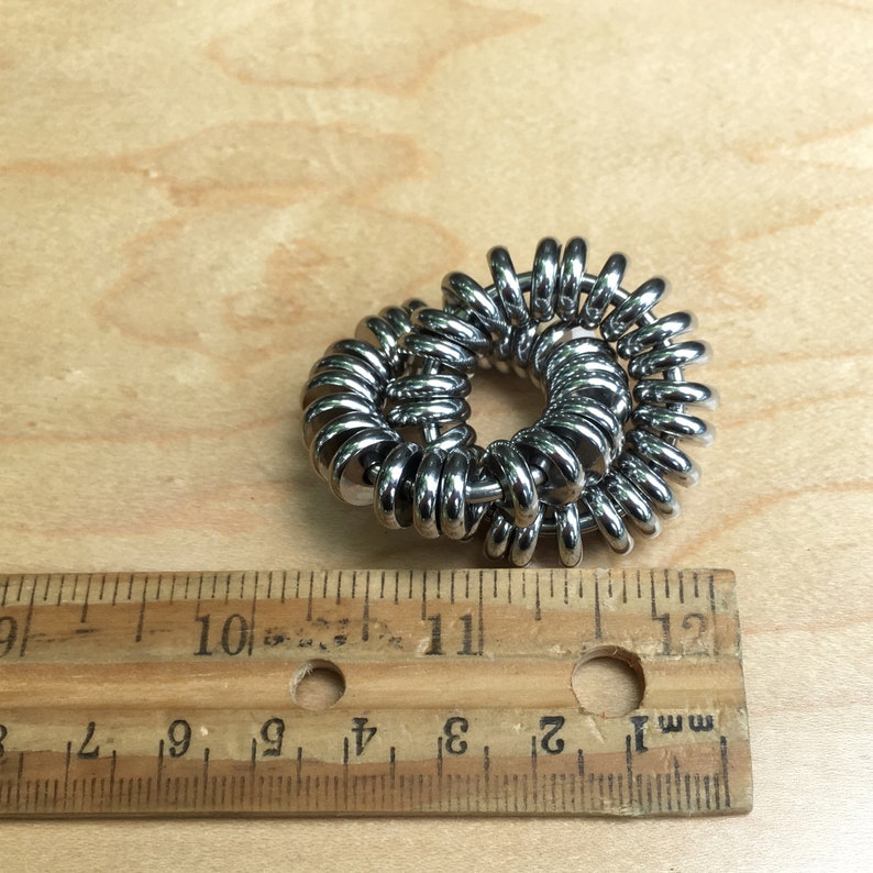 Puede incluir: Objeto met&aacute;lico plateado y brillante con forma circular, compuesto por m&uacute;ltiples anillos enrollados. El objeto tiene aproximadamente 6,35 cm de di&aacute;metro. Se encuentra sobre una superficie de madera.