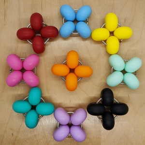 Puede incluir: Un conjunto de diez juguetes de goma redondos y coloridos, cada uno con un anillo de metal en el centro. Los juguetes est&aacute;n dispuestos en c&iacute;rculo sobre una superficie de madera.