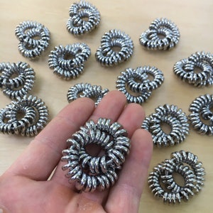 Puede incluir: Una colecci&oacute;n de coleteros plateados en forma de espiral. Uno se sostiene en una mano, mientras que otros est&aacute;n dispersos sobre una superficie de madera clara. Los coleteros est&aacute;n hechos de un material met&aacute;lico enrollado.
