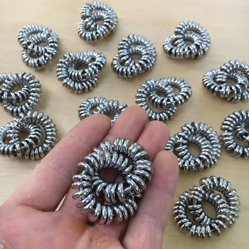 Puede incluir: Una colecci&oacute;n de coleteros plateados en forma de espiral. Uno se sostiene en una mano, mientras que otros est&aacute;n dispersos sobre una superficie de madera clara. Los coleteros est&aacute;n hechos de un material met&aacute;lico enrollado.