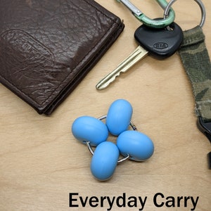 Puede incluir: Una cartera de cuero marr&oacute;n, una llave con un llavero Kia negro, un mosquet&oacute;n verde, un cord&oacute;n de camuflaje y un juguete fidget azul sobre una superficie de madera. El texto "Everyday Carry" est&aacute; escrito debajo de los objetos.
