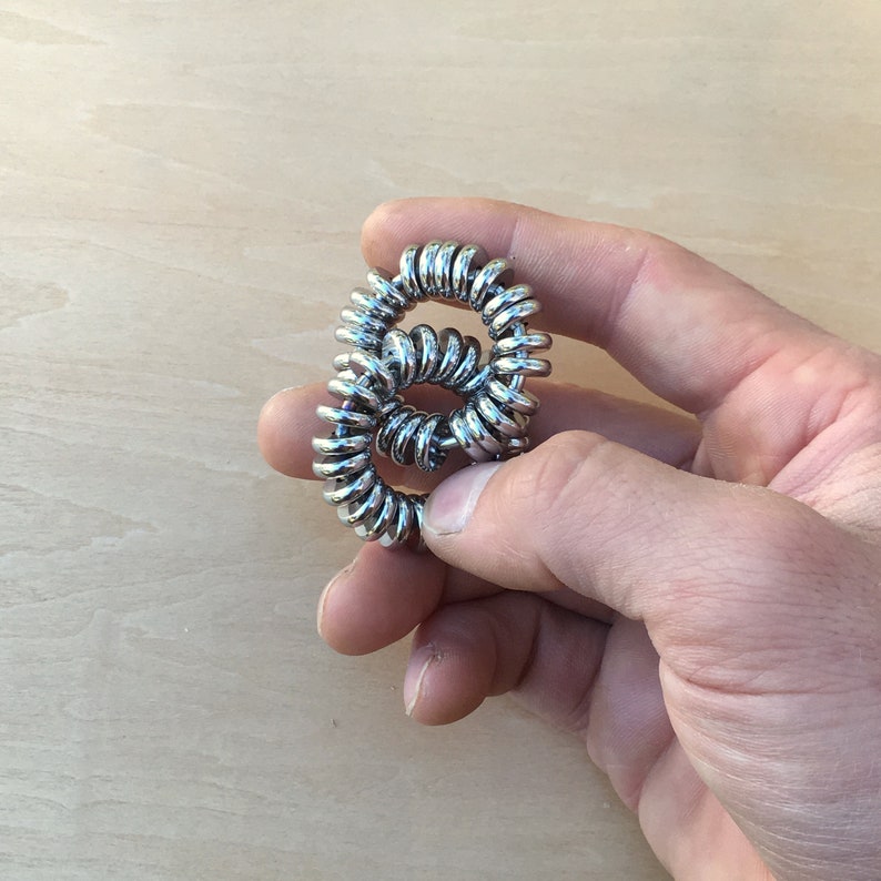 Puede incluir: Un juguete fidget de metal plateado sostenido en una mano. El juguete est&aacute; hecho de anillos de metal enrollados, formando una forma de ocho. La superficie met&aacute;lica brillante refleja la luz. El fondo es una superficie de madera de color claro.