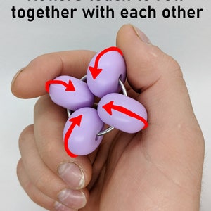 Puede incluir: Un juguete antiestr&eacute;s morado con cuatro rodillos que giran alrededor de un anillo de metal. Los rodillos est&aacute;n conectados al anillo y se tocan entre s&iacute;. El juguete se sostiene en una mano, y las flechas indican la direcci&oacute;n en la que los rodillos pueden girar.