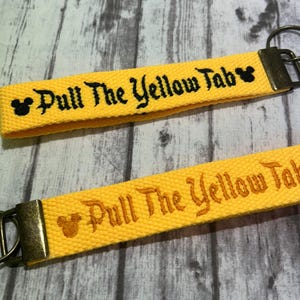 May include: Two yellow keychains with the text "Pull The Yellow Tab" in black and gold. Each keychain has a metal ring and a small Mickey Mouse head icon. The keychains are on a wooden surface.
