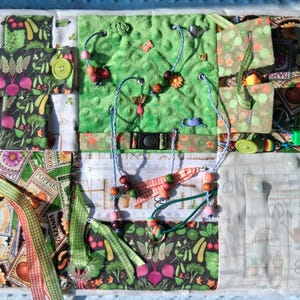 Könnte beinhalten: Ein farbenfrohes sensorisches Aktivitätsboard mit verschiedenen Texturen und interaktiven Elementen. Es zeigt Stoffquadrate mit Gemüse- und Blumenmustern, Knöpfen, Perlen, Bändern und kleinen Spielzeugen. Entwickelt für taktile Erkundung und die Entwicklung der Feinmotorik.