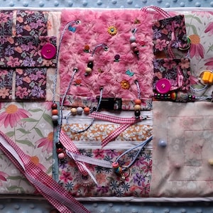 May include: A tactile activity board with diverse textures and interactive features. Includes floral fabrics, a pink plush section, buttons, beads, ribbons, and a buckle. Designed for sensory exploration and fine motor skill development.