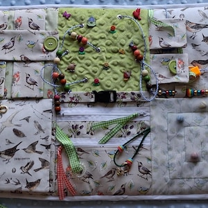 May include: A sensory activity board with various interactive elements. Features fabric with bird illustrations, pockets, zippers, buttons, beads, and a buckle. Includes green, red, and brown accents. Designed for tactile exploration and fine motor skill development.