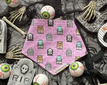 May include: A triangular pet bandana in light purple featuring a repeating pattern of cartoon monsters including vampires, mummies, and Frankenstein-like figures, with the text "LITTLE MONSTER" and a small white tag labeled "UPPERCRUFTS".