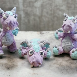 Puede incluir: Tres peluches de dragón de crochet de color morado y turquesa. Un dragón está de pie, otro está sentado y otro está acostado.