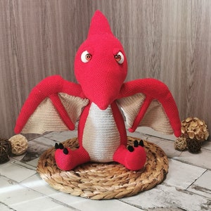 May include: A handmade red crochet pterodactyl plush toy with beige wings and black claws. The dinosaur has large, round eyes and is sitting on a woven mat. The toy is a unique, handcrafted item.