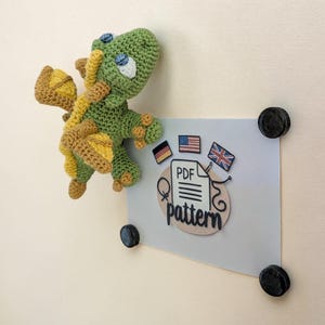 May include: A handmade green and yellow crocheted dragon with blue eyes, attached to a gray paper held by black magnets. The paper has a "PDF pattern" graphic with flags of the USA, UK, and Germany.