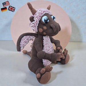 Pode incluir: Um dragão de crochê feito à mão em tons de castanho e rosa. O dragão tem olhos grandes azuis, um gorro com padrão rosa e branco e asas a condizer. O dragão está sentado com as pernas estendidas e os braços apoiados nos joelhos. O fundo é um círculo rosa claro.