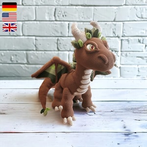 Puede incluir: Un dragón de ganchillo marrón con detalles verdes y beige. El dragón tiene ojos grandes y expresivos, cuernos beige y detalles verdes en forma de hojas. Las alas son marrones con detalles verdes y beige. El dragón está sobre una superficie de madera blanca.