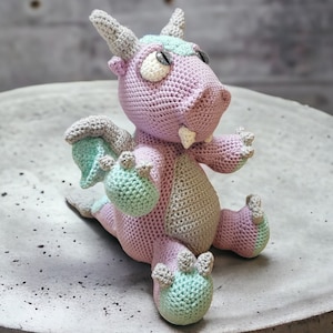 Puede incluir: Un juguete de dragón de crochet morado y verde con ojos grandes y una sonrisa de dientes. El dragón está sentado sobre una superficie gris.
