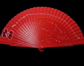 May include: A red hand fan with silver glitter and a white bow design. The fan is closed and has the word "Marisol" written on the bottom.