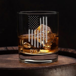 May include: A clear glass whiskey tumbler with an etched American flag design and the name "DYLAN" at the bottom. The glass is filled with amber liquid and ice cubes. The glass is sitting on a wooden barrel.