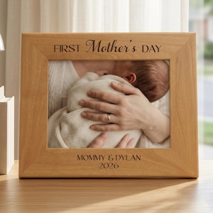 May include: A light brown wooden picture frame with the text "FIRST Mother's DAY" at the top and "MOMMY & DYLAN 2026" at the bottom. The frame holds a photo of a person holding a baby. The frame is on a wooden desk.