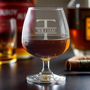 May include: A clear brandy snifter with a personalized design, filled with amber liquid. The glass features the letter "T" above the name "WILLIAM" etched into the glass. Bottles of liquor are visible in the background, along with a cigar.