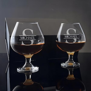 May include: Two clear brandy snifters filled with amber liquid. Each glass is engraved with the letter "S" and the name "MICHAEL". The glasses sit on a reflective black surface, with a dark background.