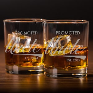 May include: Two clear glass tumblers with the text "Promoted Uncle" and "Est. 2024" etched on the front. The glasses are filled with amber liquid and ice cubes.