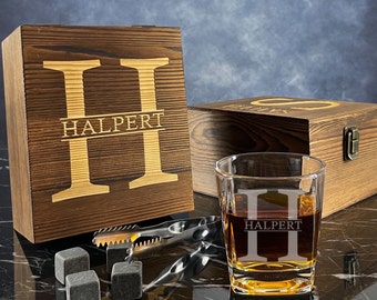 May include: A wooden box with the initials "HH" and the name "HALPERT" engraved on the front. The box contains a whiskey glass with the same initials and name engraved on it. The glass is filled with amber liquid and there are four gray whiskey stones and a pair of tongs on the table.