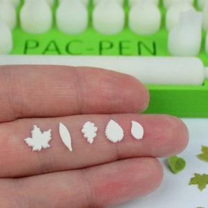 Op de afbeelding: Een hand die zes witte bladeren van polymer clay vasthoudt. De vormen omvatten een esdoornblad, een lang blad, een kleine bladcluster, een afgerond blad en twee traanvormige bladeren. De bladeren zijn in een rij op de hand gerangschikt. De achtergrond toont een groen plastic gereedschap met de tekst "PAC-PEN" erop.