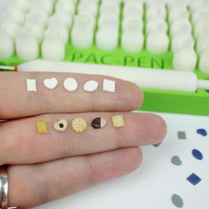 Könnte beinhalten: Miniatur-Kekse und -Formen in Weiß und Hellbraun, darunter Herzen, Quadrate und Kreise, werden auf einem Finger gezeigt. Das Wort "PAC-PEN" ist im Hintergrund zu sehen, zusammen mit einem grünen Tablett und weißen Objekten. Zusätzliche Formen sind in der Nähe verstreut.