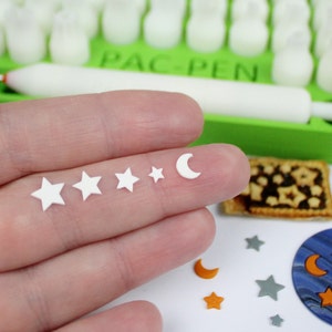 Può includere: Una mano che tiene cinque decorazioni di pasta di zucchero bianca: una luna crescente e quattro stelle di diverse dimensioni. Le decorazioni sono su una superficie bianca con altre decorazioni di pasta di zucchero, tra cui un pianeta di pasta di zucchero blu e bianco con una luna crescente e stelle, e un biscotto di pasta di zucchero marrone con un motivo a stella.