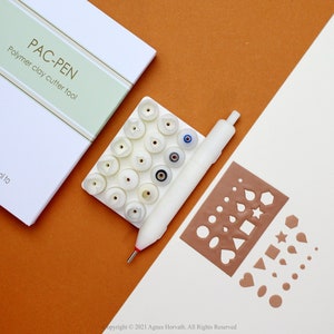 Op de afbeelding: Een witte set polymer clay cutter tools met een verscheidenheid aan verwisselbare tips. De set bevat een wit pen-achtig gereedschap met een rode punt, een witte lade met 16 verschillende tips en een bruin vel met verschillende uitgesneden vormen. De verpakking voor de gereedschapsset is wit met groene en gouden accenten en de tekst "PAC-PEN Polymer clay cutter tool".