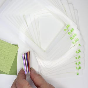May include: White, square crafting templates with green measurement markings are fanned out. A hand holds a stack of colourful paper strips. A green textured paper is also visible.