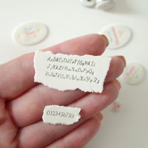 May include: Two white, irregularly shaped clay pieces with rough, torn-style edges. The larger piece features the complete alphabet in uppercase and lowercase letters, while the smaller piece displays the numbers zero through nine in a dark font.