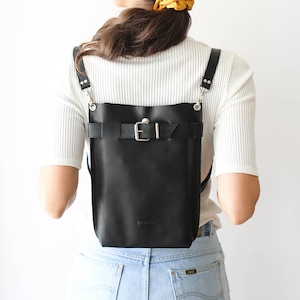 Puede incluir: Una mochila de cuero negro con una hebilla plateada y correas ajustables. La mochila la lleva puesta una persona con el pelo largo y castaño. El texto "Size Standard Convertible - Backpack / Crossbody bag" es visible en la parte superior de la imagen.