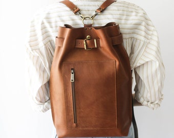 May include: A brown leather backpack with a buckle closure and a zippered pocket. The backpack has adjustable straps and is worn by a person with a white striped shirt and blue jeans.