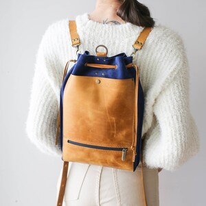 May include: A tan and navy blue leather backpack with a drawstring closure and a zippered pocket. The backpack has adjustable straps and a buckle closure.