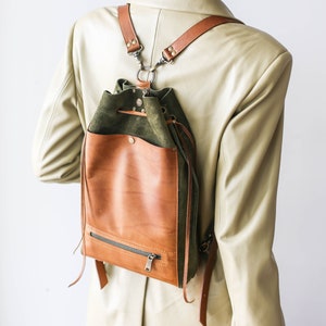 May include: Medium-sized backpack/crossbody bag in a stylish design. The bag features a combination of brown leather and olive green suede. It has brown leather straps and a zippered front pocket. The text "Medium SIZE BACKPACK/CROSSBODY STYLE" is visible.