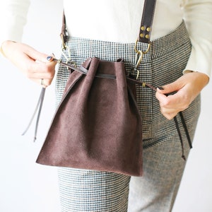 May include: A brown suede drawstring bag with a long brown leather strap. The bag is worn over the shoulder of a person wearing a white sweater and plaid pants.
