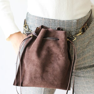 May include: A brown suede drawstring bag with a leather strap and a metal buckle. The bag is worn on a person's waist and is hanging down the side of their body.