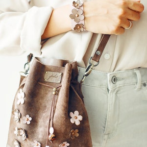 Peut inclure: Un sac seau en daim marron avec une fermeture &agrave; cordon et une bandouli&egrave;re en cuir rose. Le sac est d&eacute;cor&eacute; de fleurs en cuir rose et porte une petite &eacute;tiquette en cuir marron avec le mot "feels" dessus.