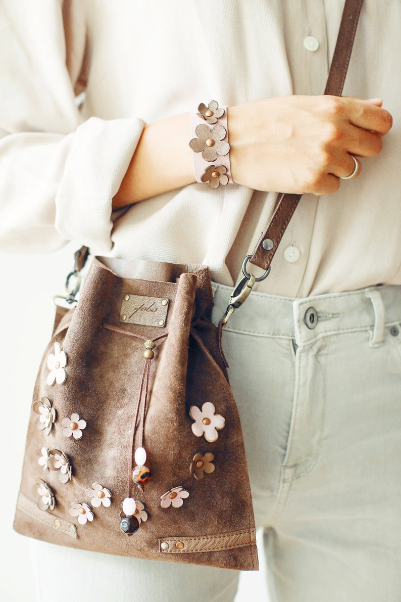 Peut inclure: Un sac seau en daim marron avec une fermeture &agrave; cordon et une bandouli&egrave;re en cuir rose. Le sac est d&eacute;cor&eacute; de fleurs en cuir rose et porte une petite &eacute;tiquette en cuir marron avec le mot "feels" dessus.
