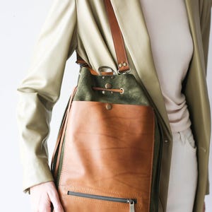 May include: A two-tone leather and suede shoulder bag. The bag features a brown leather front pocket with a zipper, a green suede upper section, and brown leather straps. The bag is worn over the shoulder.