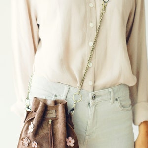 Peut inclure: Un sac seau en daim marron avec une longue bandouli&egrave;re en cha&icirc;ne et des embellissements de fleurs roses. Le sac a une fermeture &agrave; cordon et un pompon.