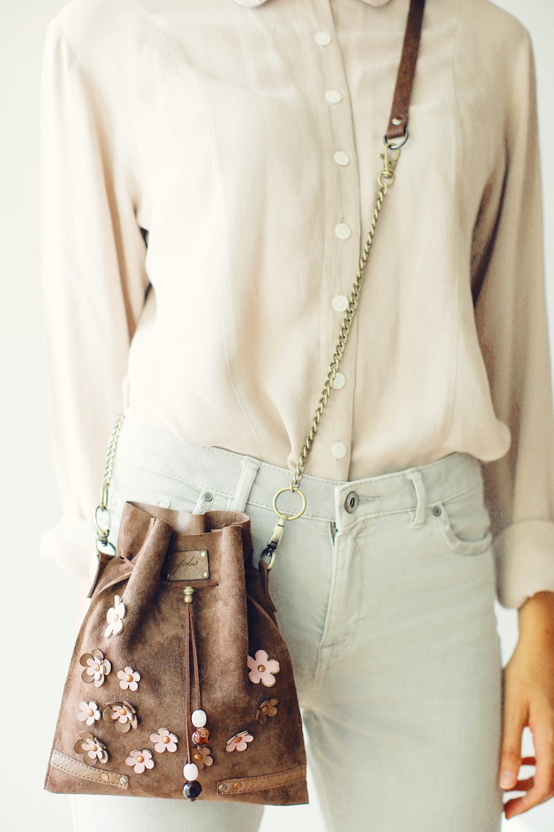 Peut inclure: Un sac seau en daim marron avec une longue bandouli&egrave;re en cha&icirc;ne et des embellissements de fleurs roses. Le sac a une fermeture &agrave; cordon et un pompon.