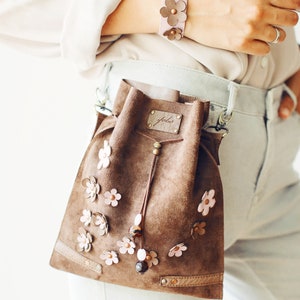 Peut inclure: Un sac seau en daim marron avec une fermeture &agrave; cordon et une petite &eacute;tiquette en cuir qui dit "Jule". Le sac est d&eacute;cor&eacute; de fleurs en cuir rose et marron et poss&egrave;de une bandouli&egrave;re en cuir marron.