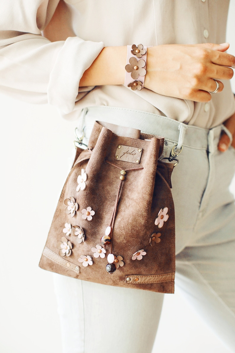 Peut inclure: Un sac seau en daim marron avec une fermeture &agrave; cordon et une petite &eacute;tiquette en cuir qui dit "Jule". Le sac est d&eacute;cor&eacute; de fleurs en cuir rose et marron et poss&egrave;de une bandouli&egrave;re en cuir marron.