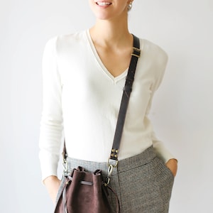 May include: A woman wearing a white sweater and plaid pants is carrying a brown leather bucket bag with a long strap.