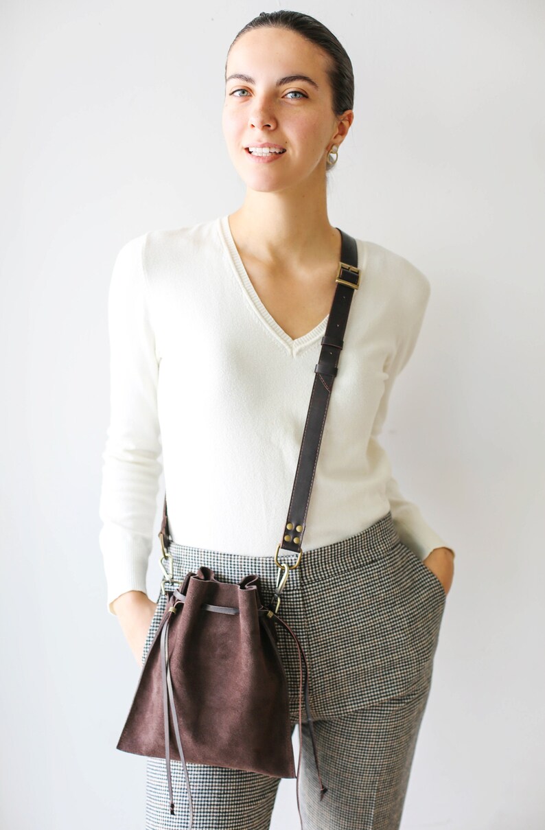 May include: A woman wearing a white sweater and plaid pants is carrying a brown leather bucket bag with a long strap.