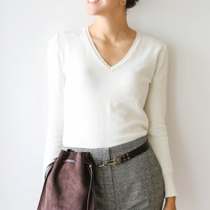 May include: A woman wearing a white v-neck sweater and black and white checkered pants with a brown suede bag attached to a brown leather belt.