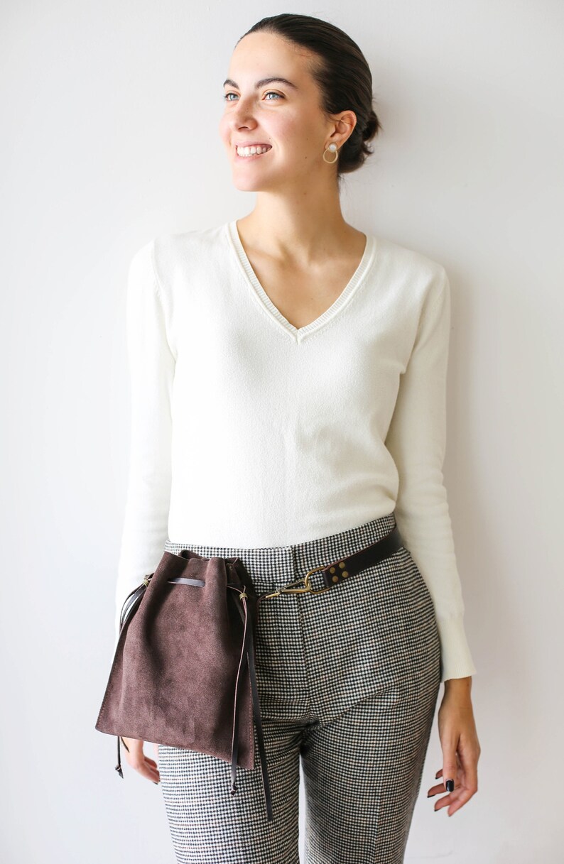 May include: A woman wearing a white v-neck sweater and black and white checkered pants with a brown suede bag attached to a brown leather belt.