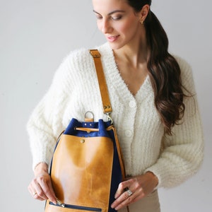 May include: A tan leather bucket bag with a navy blue suede bottom. The bag has a drawstring closure and a zippered pocket on the front.